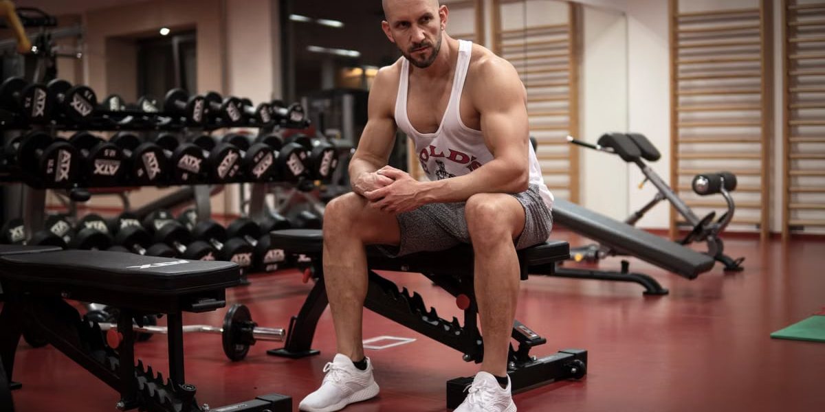 Strong, determined man in white tank top sitting in gym, showcasing fitness and strength.