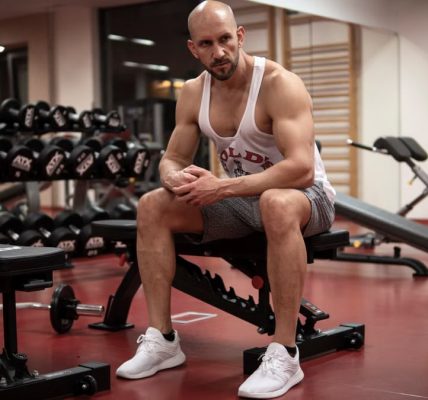 Strong, determined man in white tank top sitting in gym, showcasing fitness and strength.