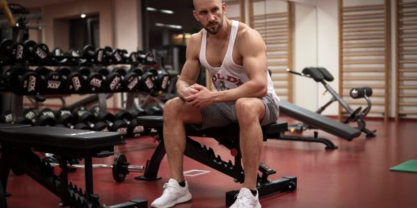 Strong, determined man in white tank top sitting in gym, showcasing fitness and strength.